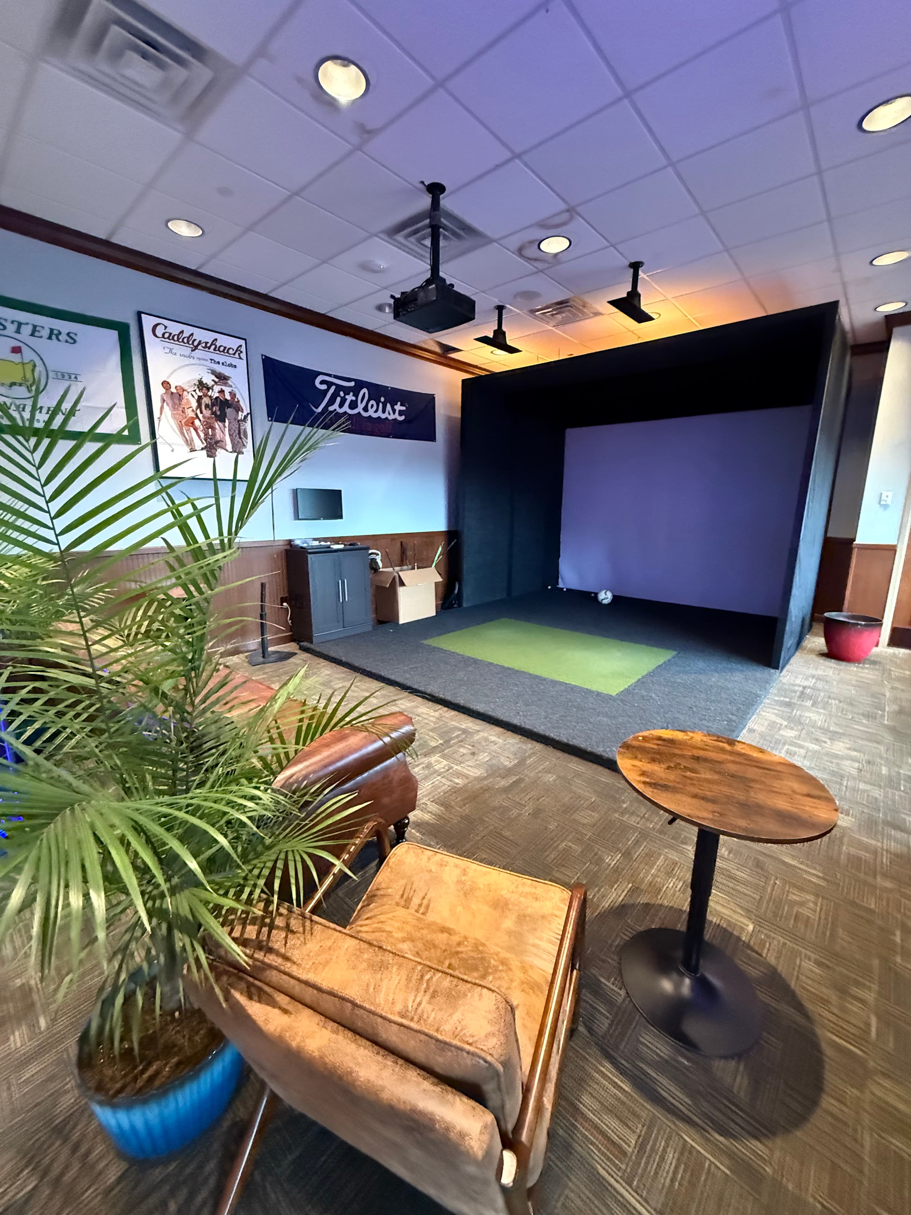 Golf simulator room with Titleist branding, lounge seating, and tropical plants